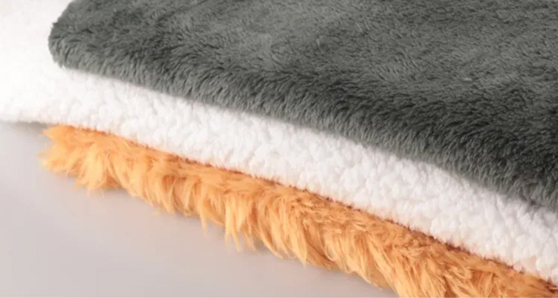 Stacked plush fabrics in gray, white, and orange, showing different pile lengths and textures commonly used for stuffed animals and plush toys.