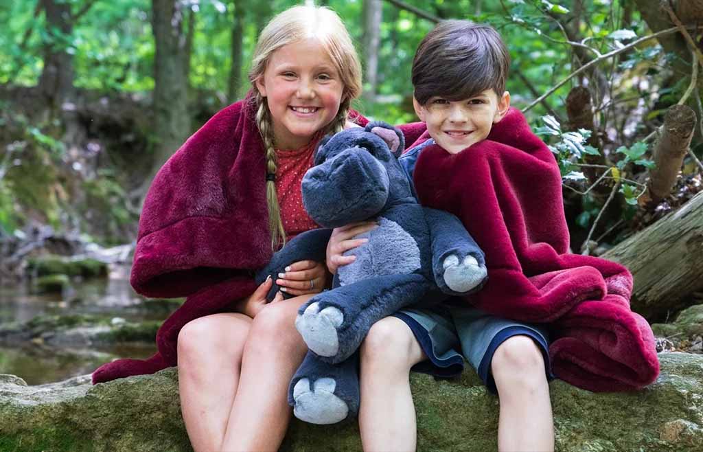 Two smiling children wrapped in soft blankets holding a plush hippo toy while sitting in a forest setting.