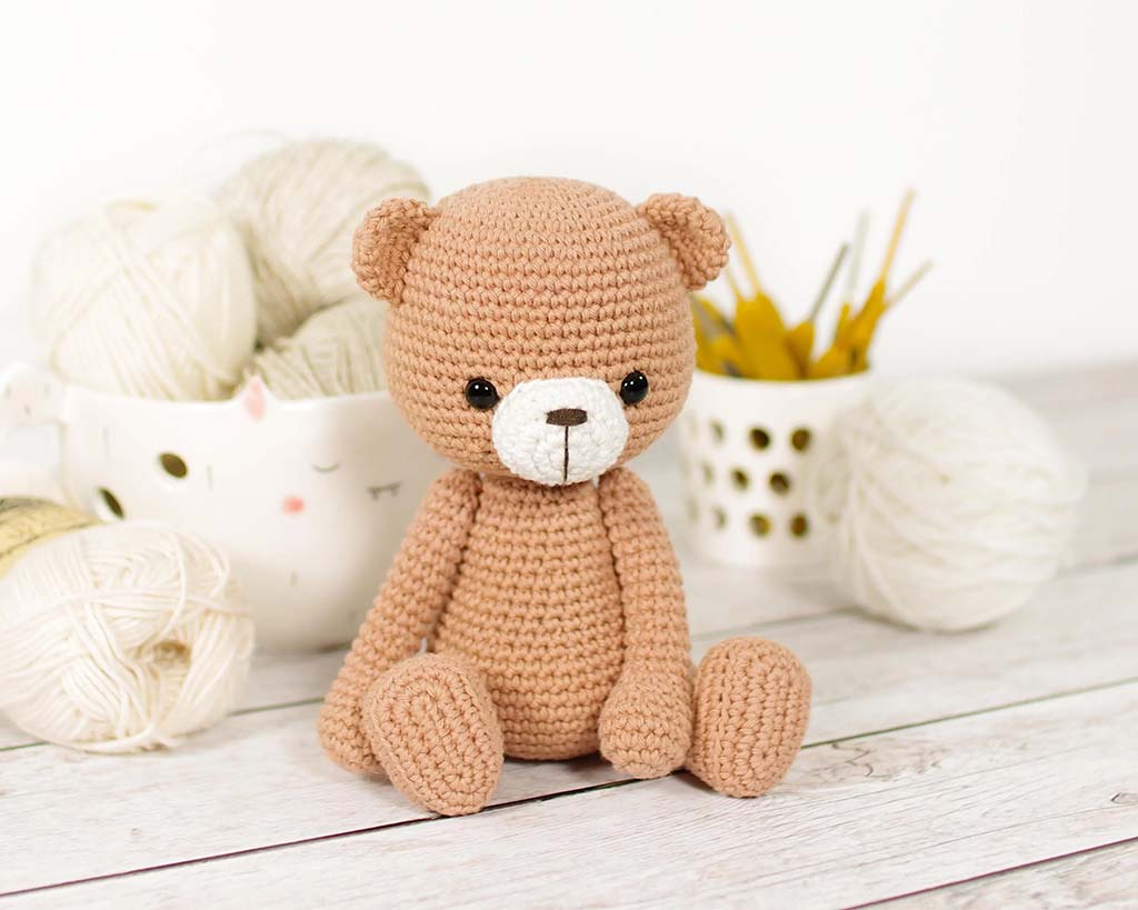 Cute handmade crochet teddy bear sitting on a wooden table with yarn balls and crochet hooks in the background.