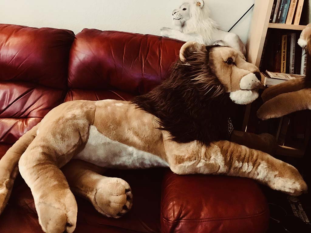 Oversized plush lion toy with a fluffy mane lying on a red leather sofa, with a smaller white lion plush placed on the backrest.