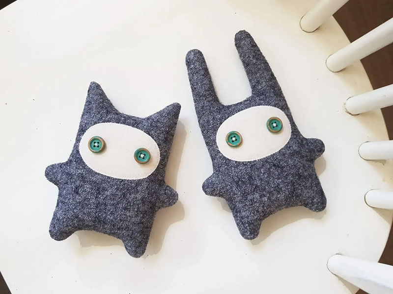 Two handmade plush creatures made from dark blue textured fabric, each with a white oval face and green button eyes, placed on a white surface.