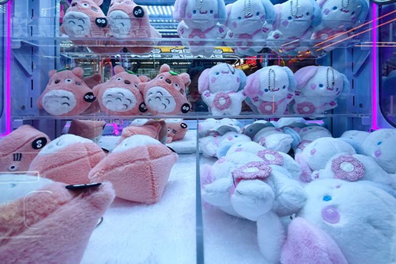 A claw machine filled with pastel pink and white plush toys, including round animals and bunny characters, arranged on upper shelves and the bottom platform under blue and purple neon lights.