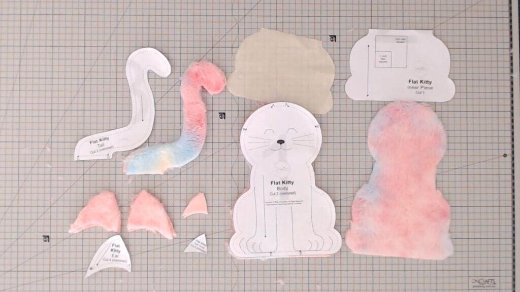 Flat Kitty plush sewing pattern pieces laid out on a gridded cutting mat, including body, tail, ears, inner piece, and cut fabric sections in pastel faux fur and beige lining.