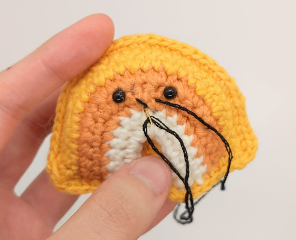 Close-up of a hand embroidering facial details on a small yellow and orange crocheted amigurumi piece using black yarn, with safety eyes already attached.