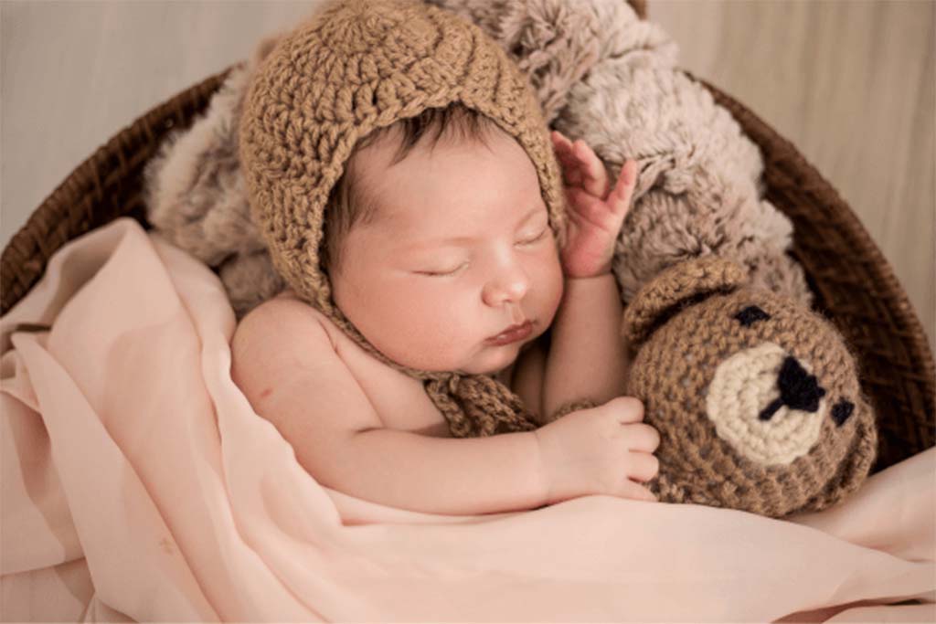 A newborn baby sleeping in a basket while holding a knitted teddy bear and wearing a crochet bonnet.