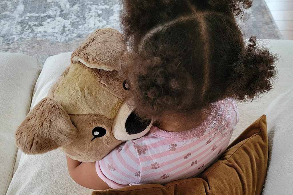 A young child with curly hair, seen from behind, hugging a large soft plush bear while sitting on a couch.