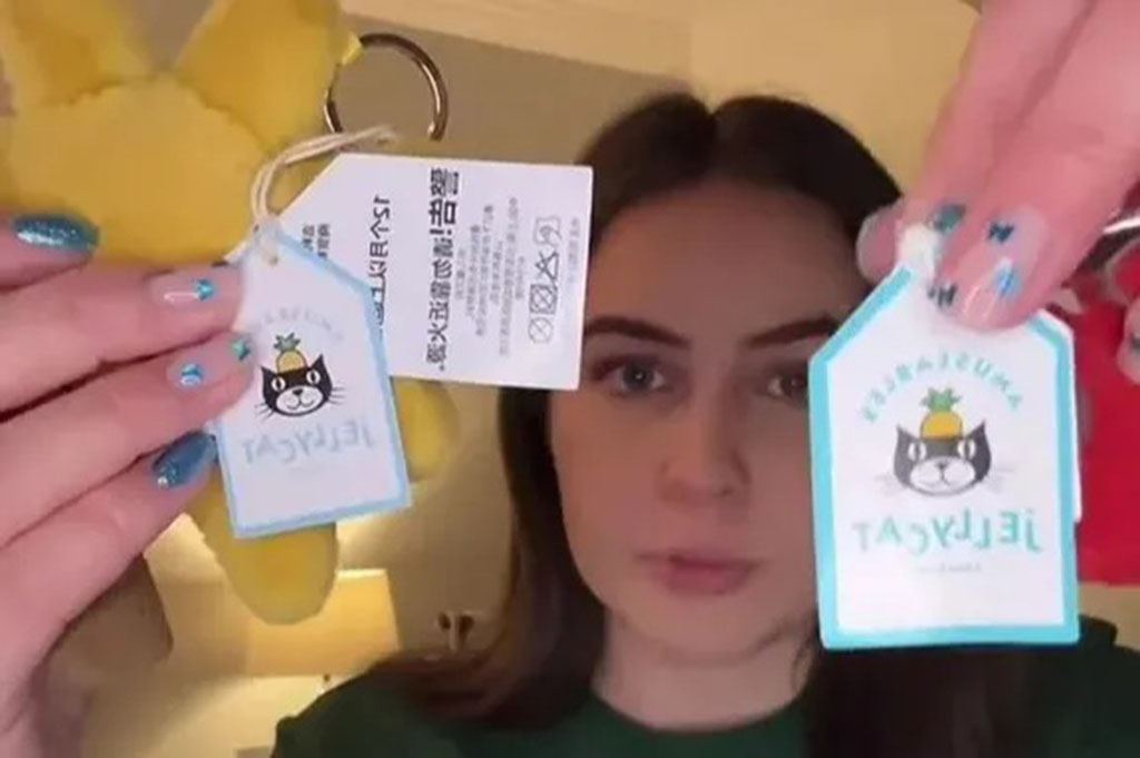 A woman holds up two Jellycat-style product tags side by side, showing differences in design and printing while a plush toy hangs behind them, highlighting a comparison between tags.