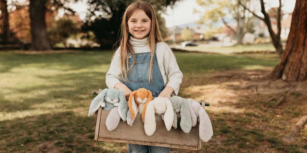 Smiling girl in denim overalls holding a wooden box filled with soft bunny plush toys in various pastel colors in a sunny park.