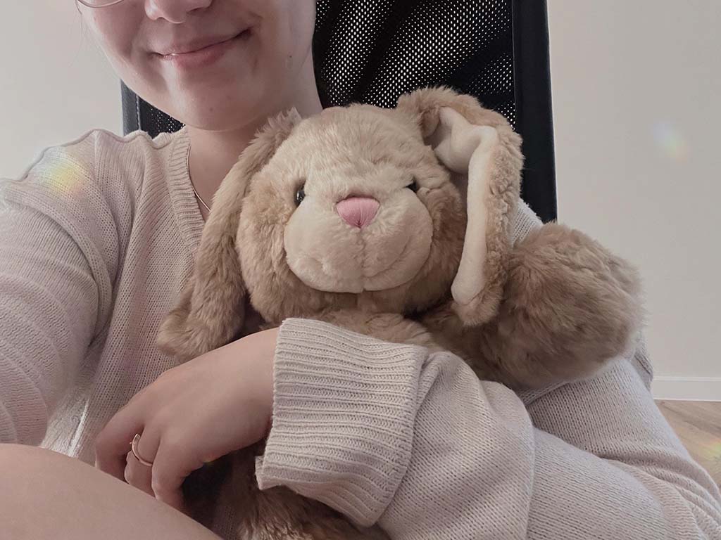A young woman gently hugging a fluffy beige bunny plush toy with floppy ears, smiling softly while sitting in a cozy indoor setting.