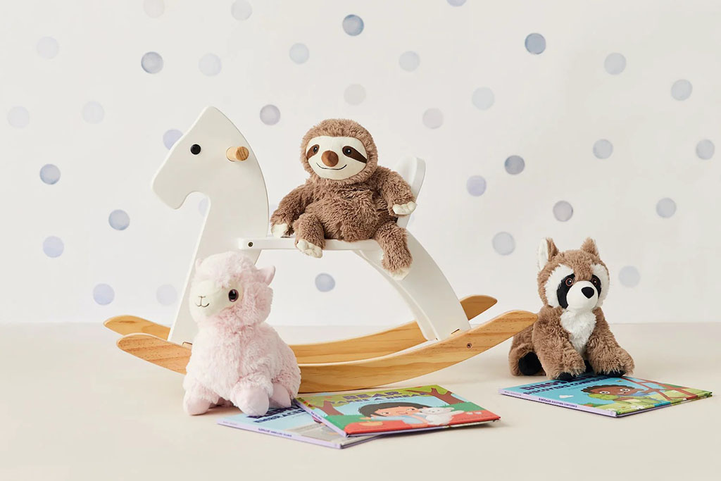 A sloth plush toy sits on a white wooden rocking horse, with a pink alpaca plush and a raccoon plush placed nearby on top of colorful children’s books in a soft, pastel nursery setting.