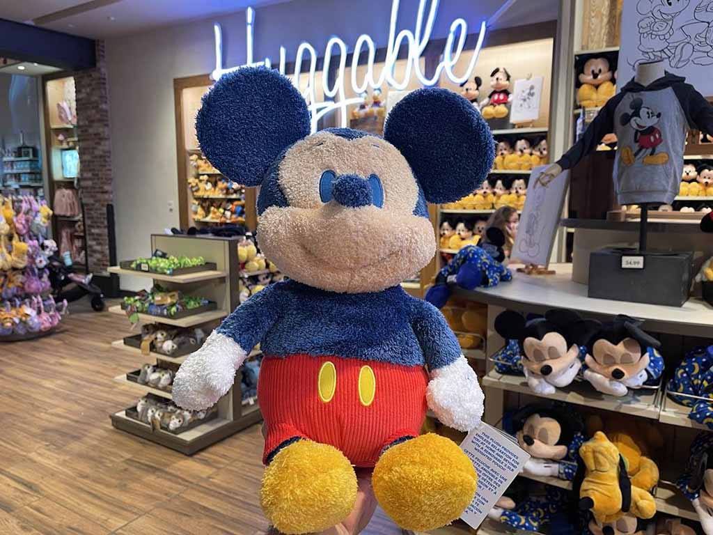 Hand holding a soft weighted Mickey Mouse plush toy inside a Disney store with “Huggable” sign and shelves full of Mickey merchandise in the background.