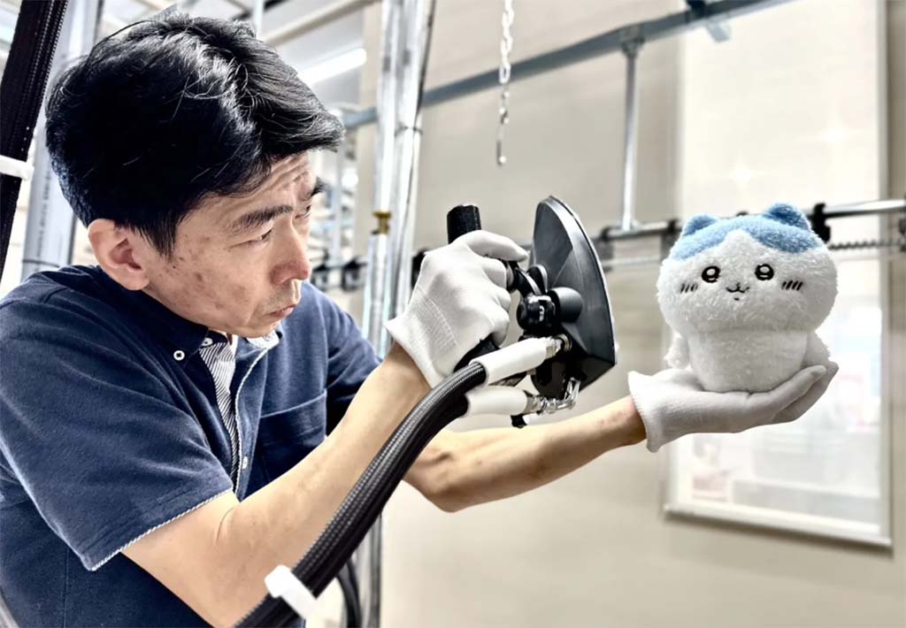 Factory worker wearing gloves carefully inspects a small white plush toy with blue ears using a handheld testing tool, ensuring plush quality control.
