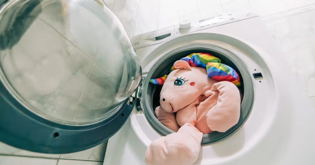 A large pink unicorn plush toy with rainbow hair placed inside a front-loading washing machine before cleaning.
