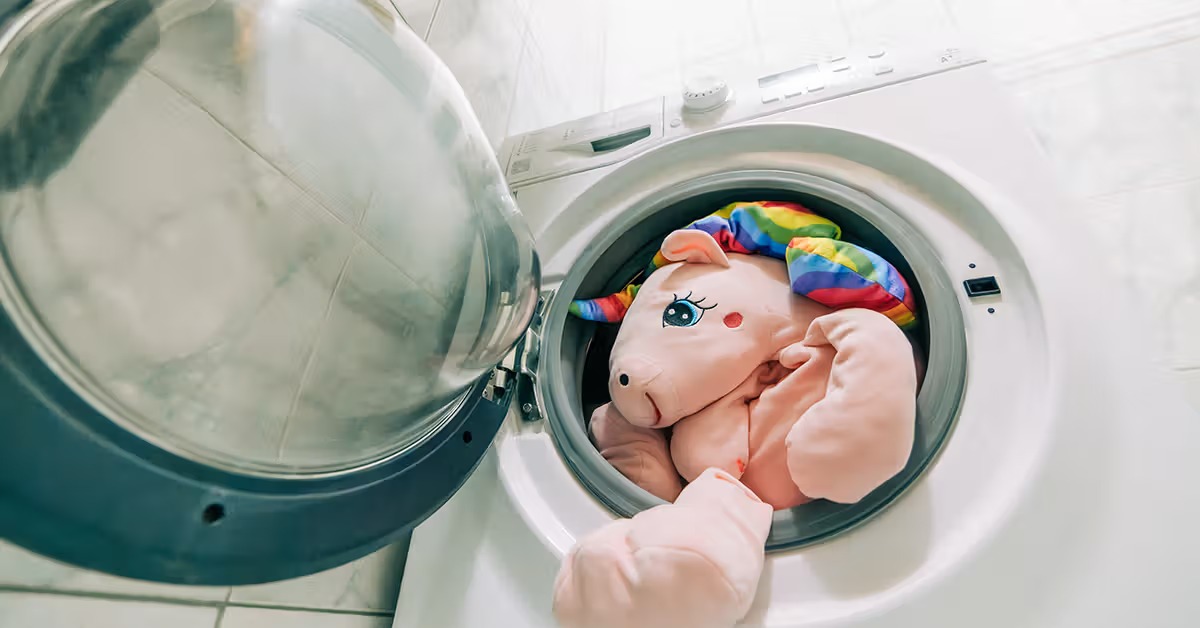 A large pink unicorn plush toy with rainbow hair placed inside a front-loading washing machine before cleaning.