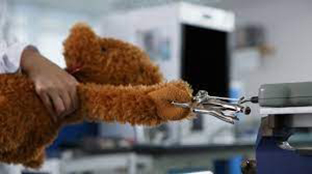 A laboratory technician performs a tensile strength test on a plush teddy bear’s arm using a mechanical testing device in a safety compliance lab.