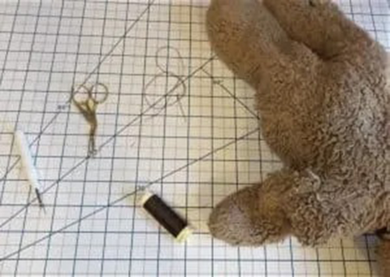 Top-down view of sewing tools—scissors, needle, thread, and seam ripper—beside a damaged plush toy on a grid cutting mat.