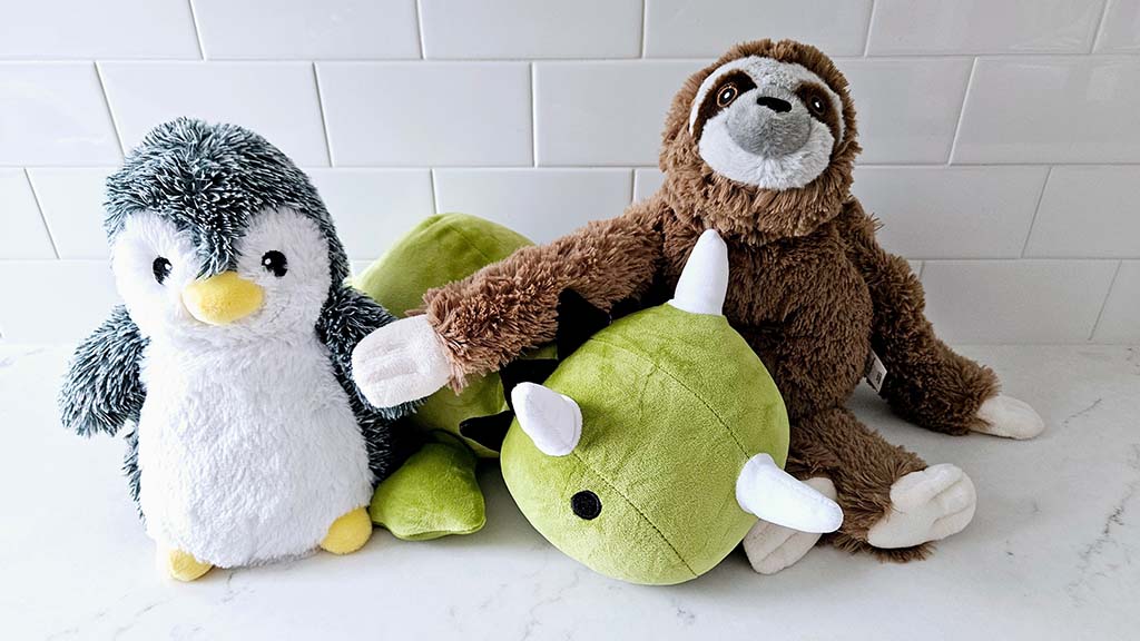 A group of plush toys including a fluffy penguin, a brown sloth, and a green dinosaur lying on a white surface with a tiled background.
