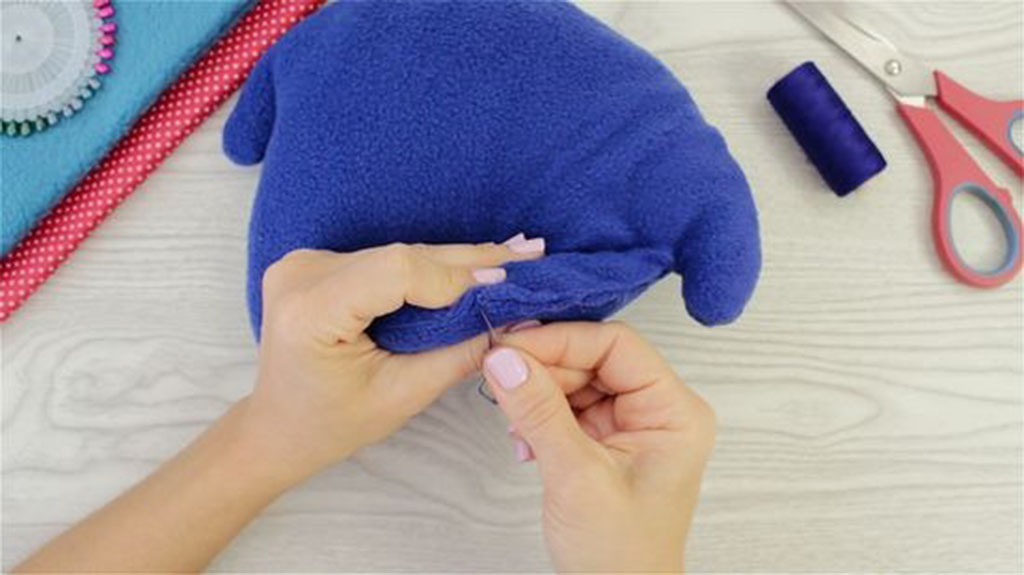 Close-up of hands sewing a blue plush toy by hand using a needle and thread, finishing seam details on a crafting table.