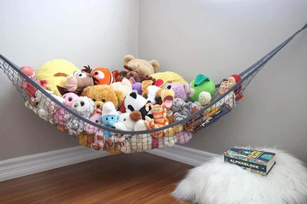 A stuffed animal hammock filled with various plush toys hanging in a corner of a room above the floor.