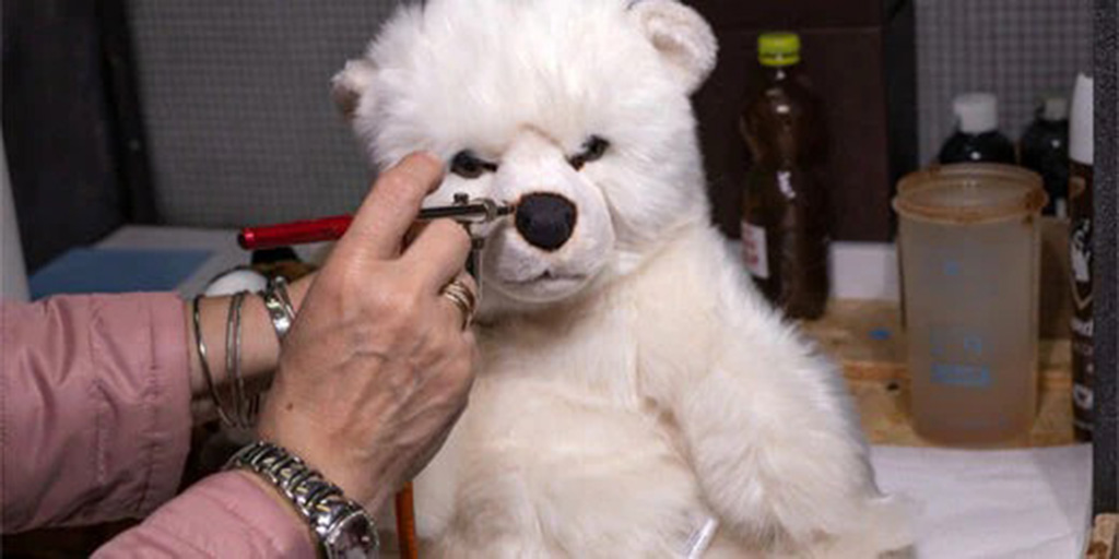 A person uses an airbrush tool to add shading to the nose of a white plush bear, showing part of the toy-painting process on a worktable with bottles and tools in the background.
