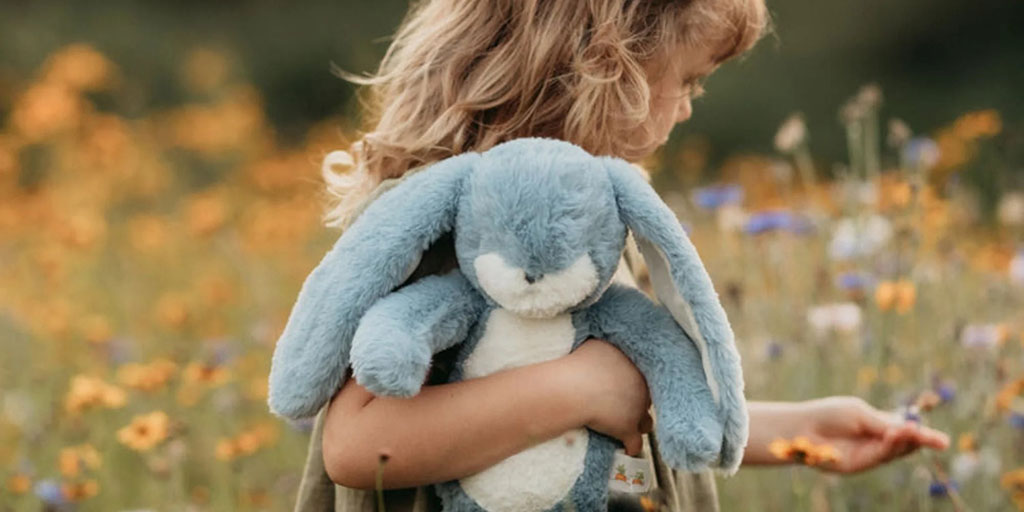 Young girl holding a soft blue bunny plush toy while standing in a field of wildflowers.