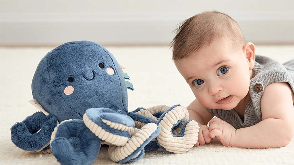 A baby lying on the floor beside a soft blue octopus plush toy with rope-style tentacles, looking forward with wide eyes.