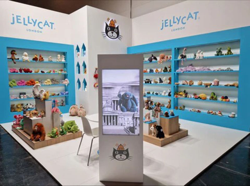 A Jellycat trade show booth featuring shelves filled with plush toys, a central display screen, and bright blue-and-white branded walls showcasing the London-based plush company.