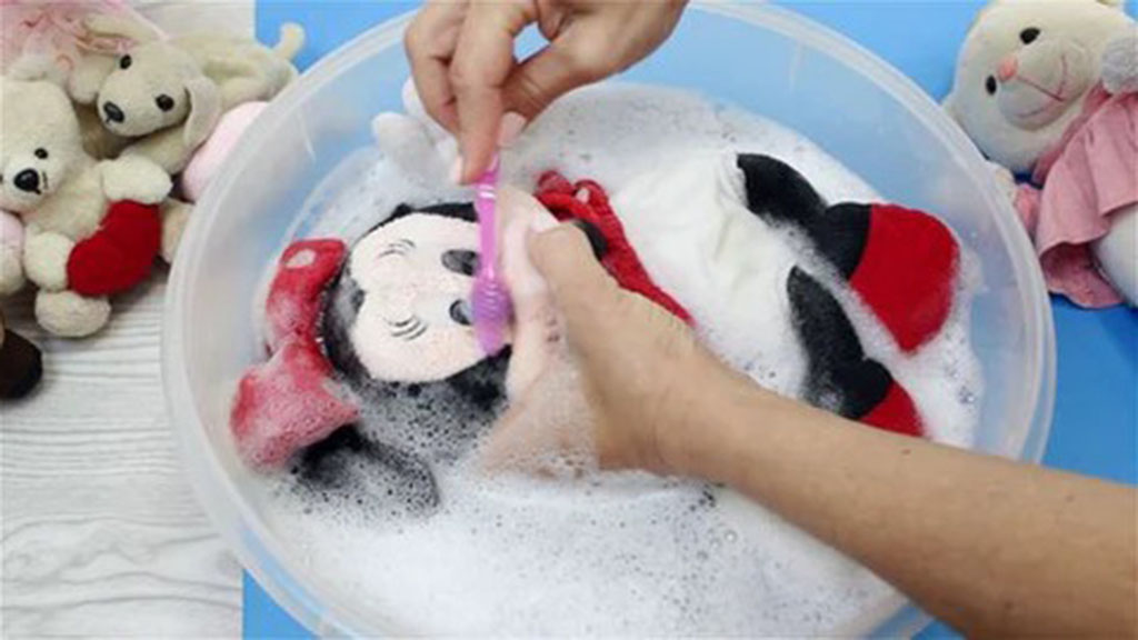 Hands cleaning a plush doll in a basin filled with soapy water using a soft brush, with other stuffed animals placed nearby.