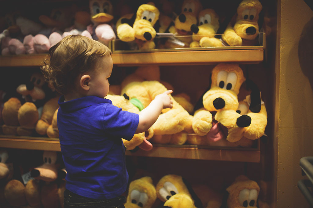 Young child in a blue shirt pointing at a shelf full of yellow Pluto plush toys in a Disney store.