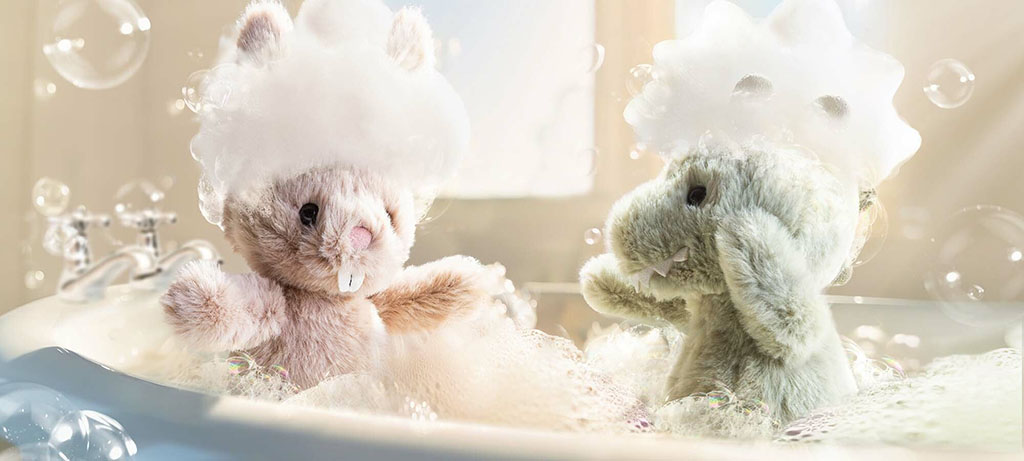 Two plush toys—a bunny and a dinosaur—sit together in a bubbly bathtub, each wearing a foam bubble hat, surrounded by bright light and playful soap bubbles.