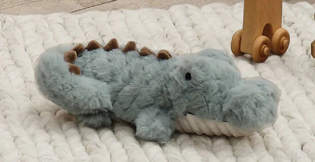Light blue crocodile plush toy with brown spikes lying on a soft white knitted rug near wooden toys.