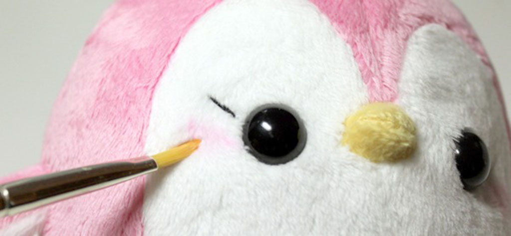 Hand applying blush with a small brush onto the cheek of a pink and white plush penguin.