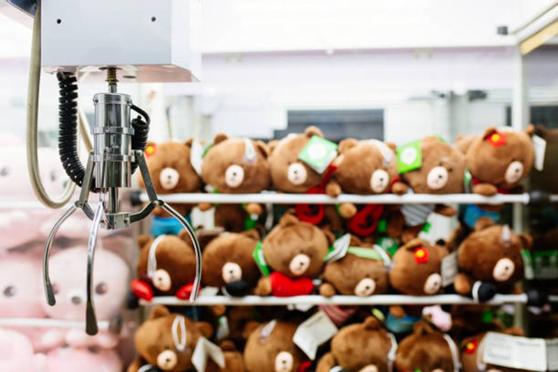 A claw machine with a metal grabbing claw in the foreground, positioned above shelves filled with brown teddy bear plush toys wearing colorful accessories inside a brightly lit arcade.