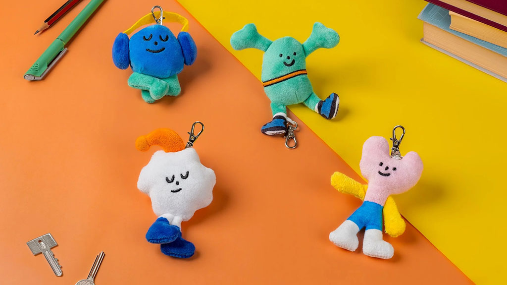 Four colorful plush character keychains arranged on a two-tone orange and yellow background with stationery and books nearby.