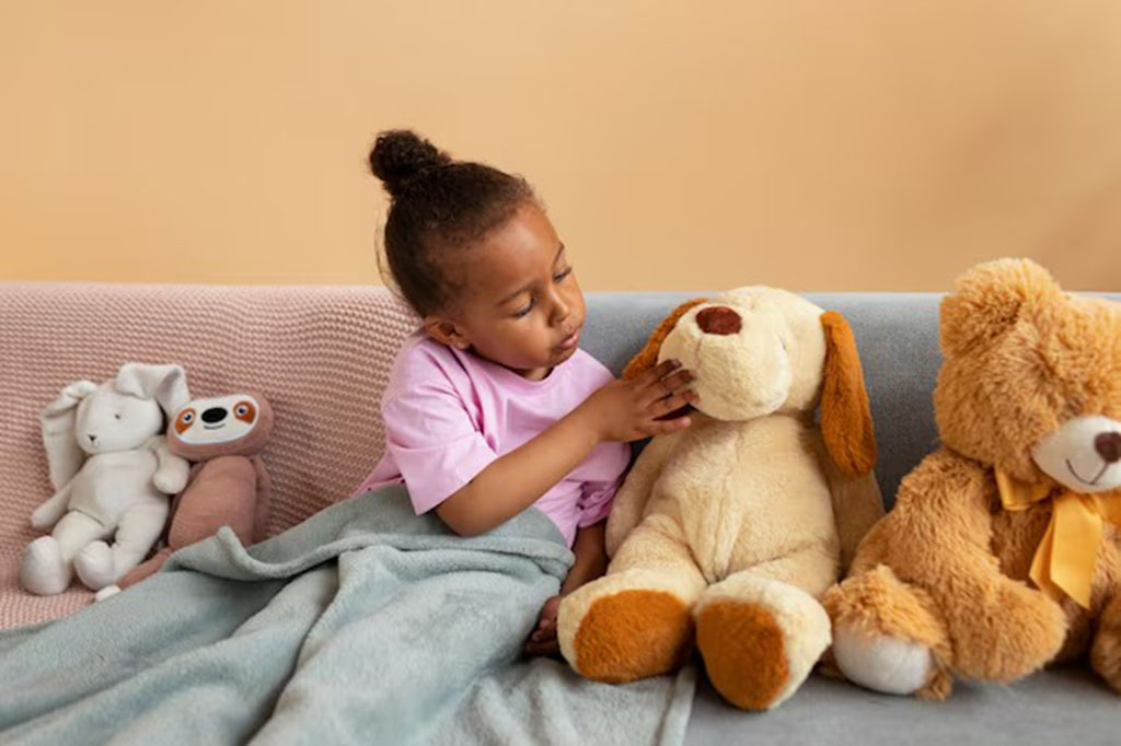 A young child sitting on a sofa under a blanket, gently touching a large plush dog, surrounded by several stuffed animals including a white bunny, a pink toy, and a teddy bear.