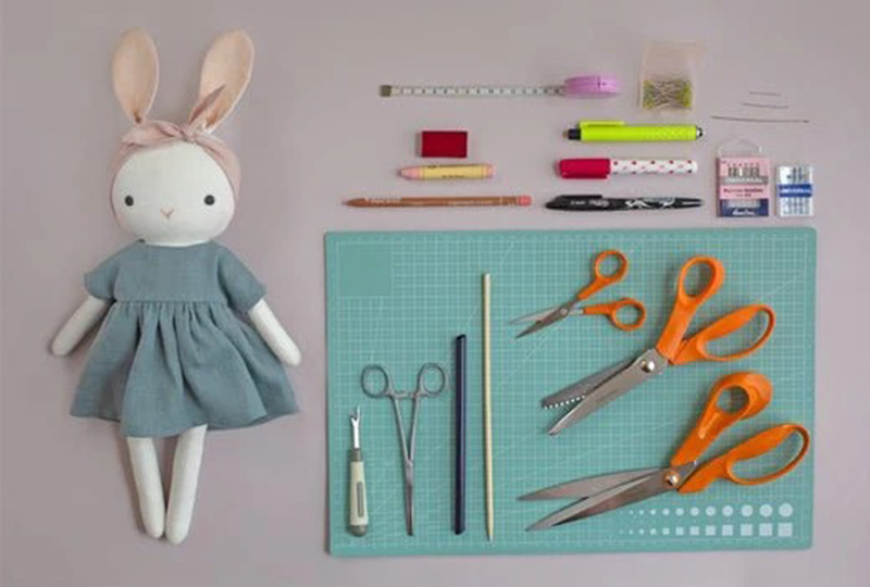 Flat lay of doll sewing tools and handmade cloth bunny doll on a crafting mat.