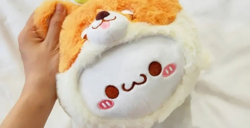 A hand holding a round plush toy featuring a smiling face with blushing cheeks and an orange animal hood design, placed on a white background.