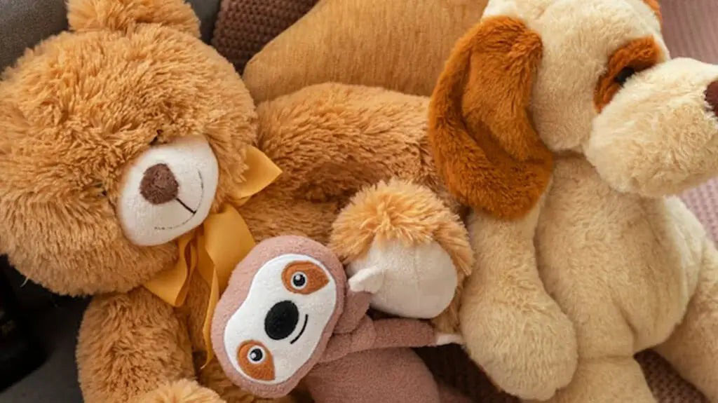 A cozy group of plush toys featuring a large teddy bear with a yellow ribbon, a soft brown dog, and a small smiling sloth doll nestled between them.