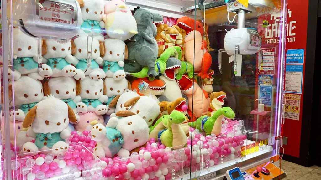 A claw machine filled with stacked plush dogs on the left and colorful dinosaur plush toys on the right, surrounded by pink and white plastic balls.