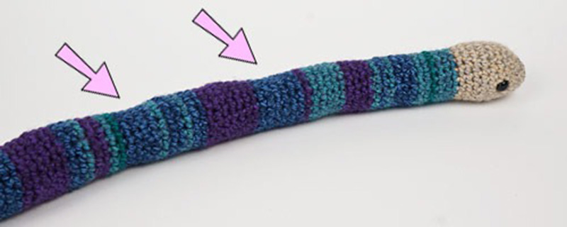 A long crocheted snake toy with visible uneven stuffing areas highlighted by arrows.