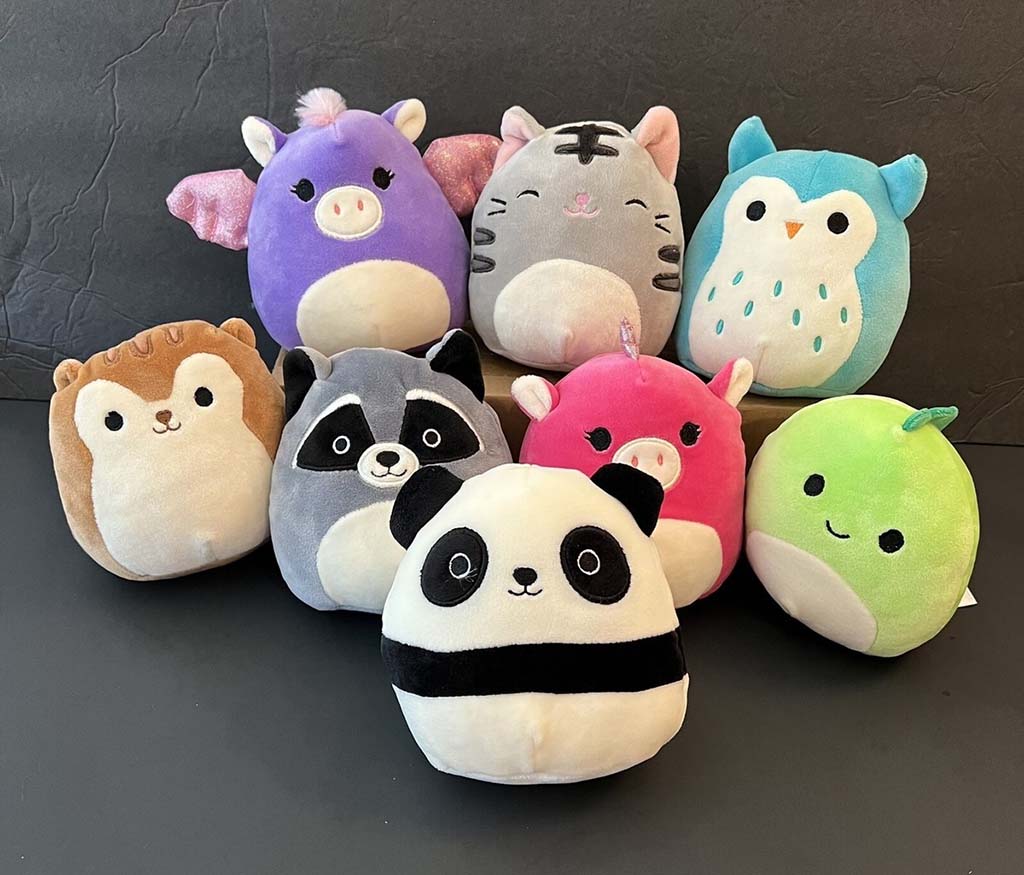 A colorful collection of Squishmallow-style animal plush toys, including panda, raccoon, hedgehog, unicorn, cat, pig, owl, and green fruit characters arranged together on a dark background.