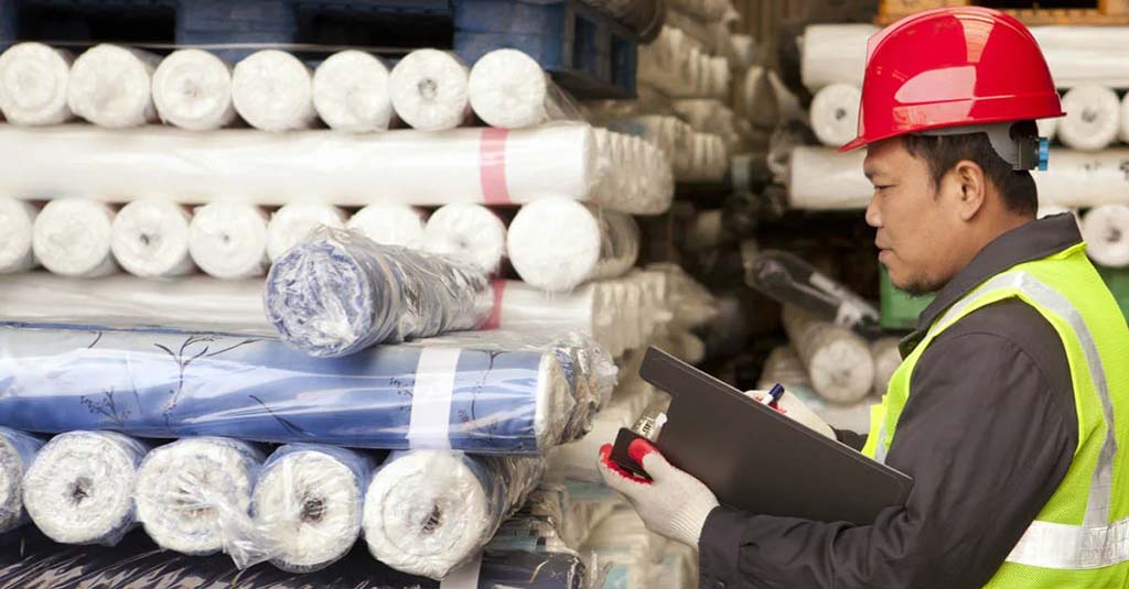 Worker in a red safety helmet inspecting and recording inventory of rolled fabric materials in a warehouse, representing quality control and material management in manufacturing.