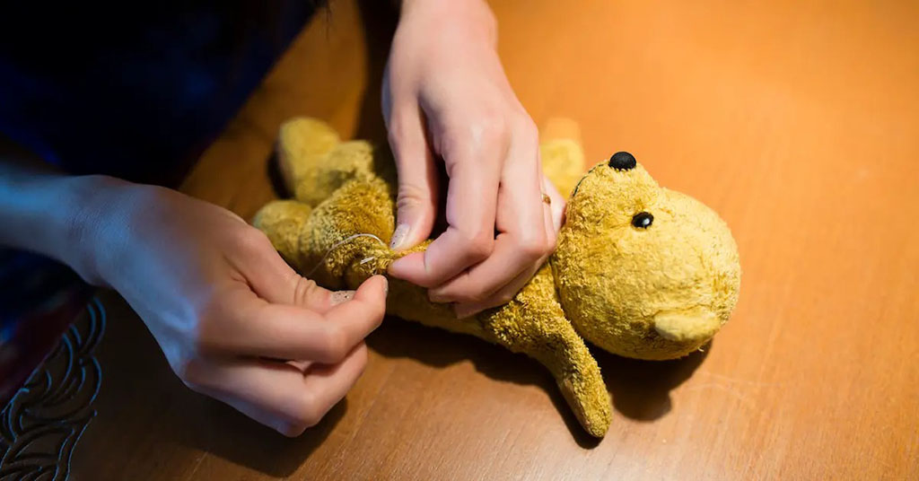 Hands sewing and repairing a worn yellow teddy bear on a wooden table.