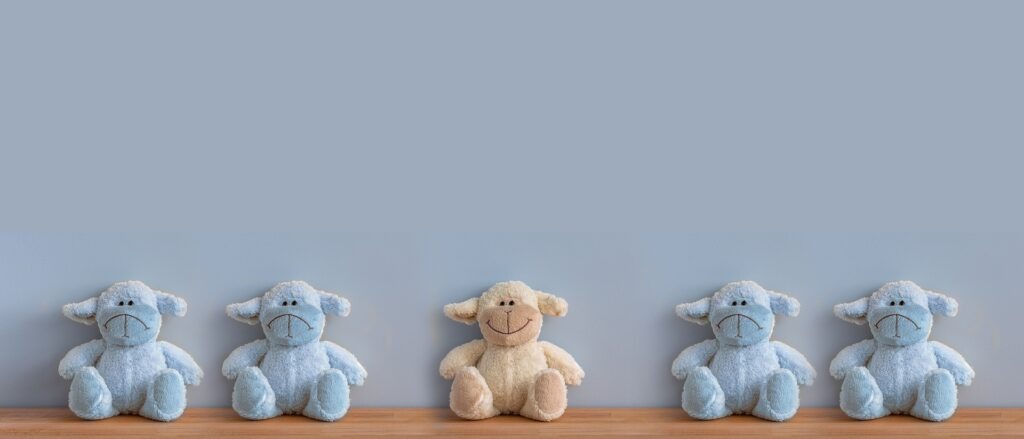 Row of small plush sheep toys on a wooden surface, with one beige smiling sheep in the center surrounded by blue sheep with neutral expressions against a soft gray background.