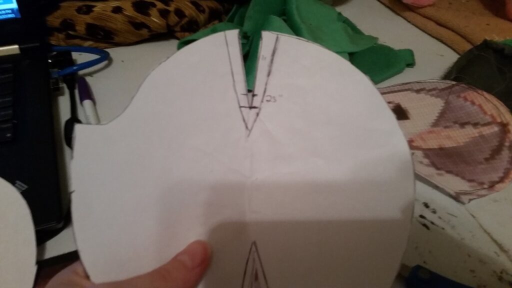 Hand holding a paper plush toy pattern piece with a drawn dart marked in pencil, surrounded by other fabric and pattern pieces on a work table