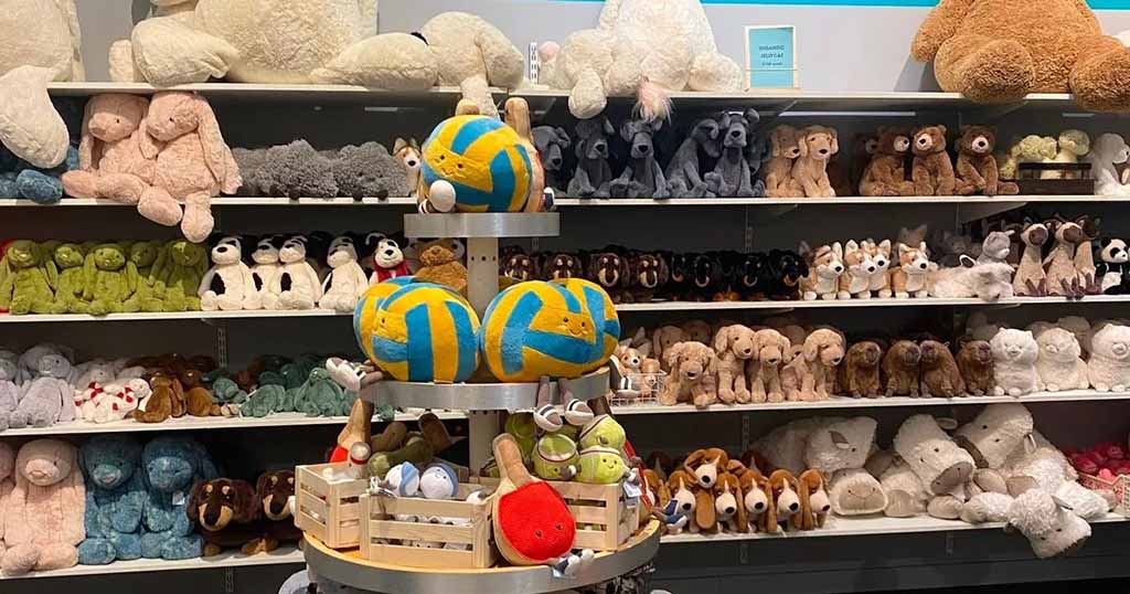 A wide shelf-packed display of plush toys arranged by color and animal type in a toy store, featuring bunnies, dogs, bears, frogs, and more, with a central round stand holding colorful plush balls and small stuffed characters in wooden crates.