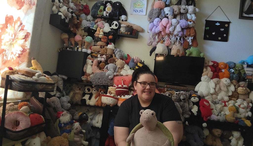 A smiling person sitting in a room filled with shelves and piles of plush toys in various colors, shapes, and sizes, showcasing an impressive stuffed animal collection.