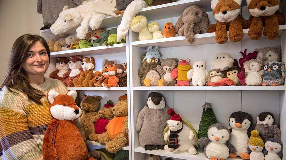 A woman standing beside shelves filled with various plush animals, including foxes, penguins, owls, bears, and dragons, while holding a large fox plush toy.