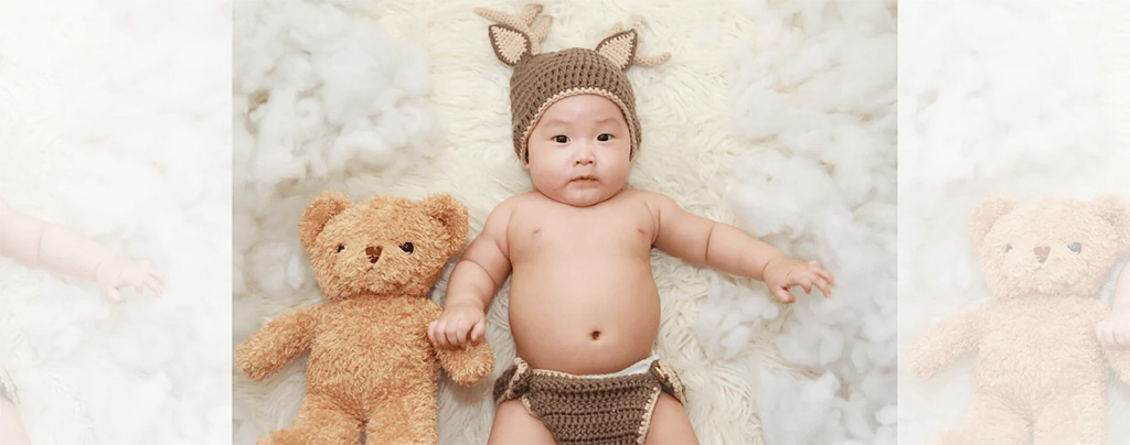 A baby wearing a knitted hat with ears lies on a fluffy cream-colored blanket, holding a brown teddy bear beside them.
