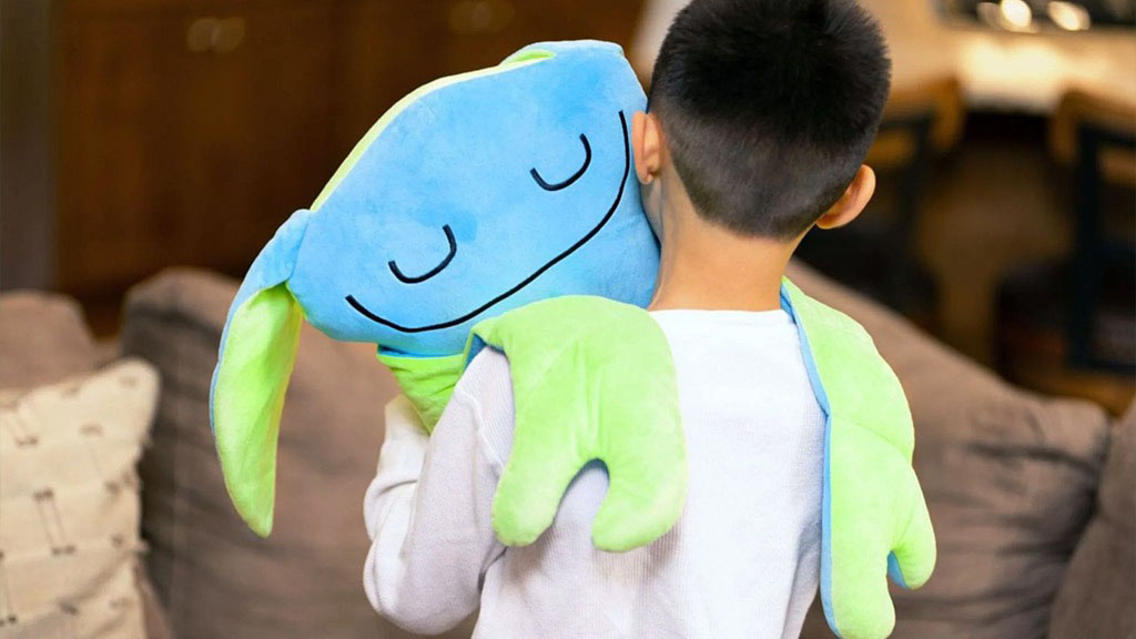 A young boy hugging a blue and green plush frog toy with a smiling face while standing in a cozy living room.